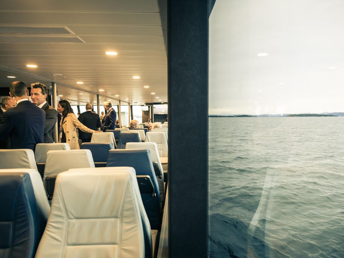 People are gathered and conversing inside a ferry with a view of the sea through large windows.